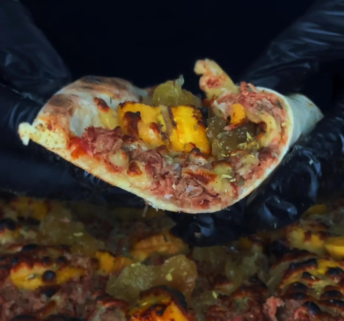 Fatia de pizza de costelinha com banana, segurada por mãos com luvas pretas sobre a pizza inteira